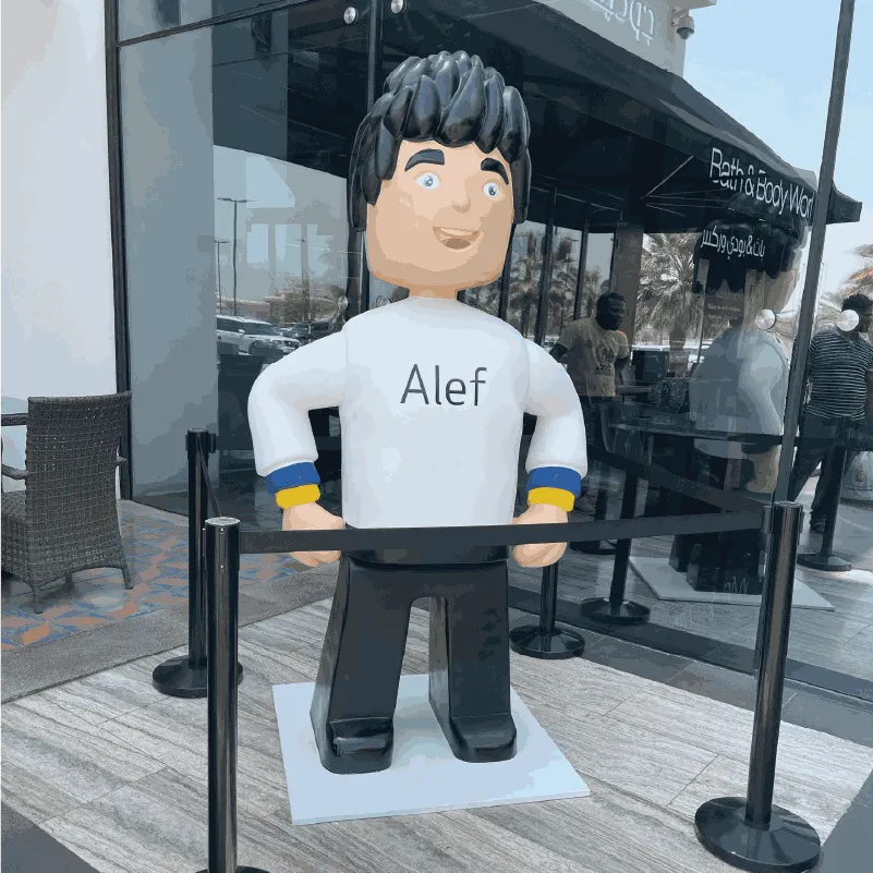 Large statue of a cartoon character with black hair wearing a white shirt labeled Alef outside a store.