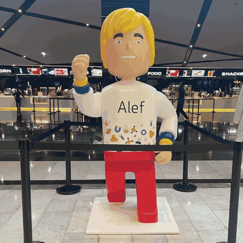 Large Lego-style figure with blond hair wearing 'Alef' shirt and red pants stands in a mall area.