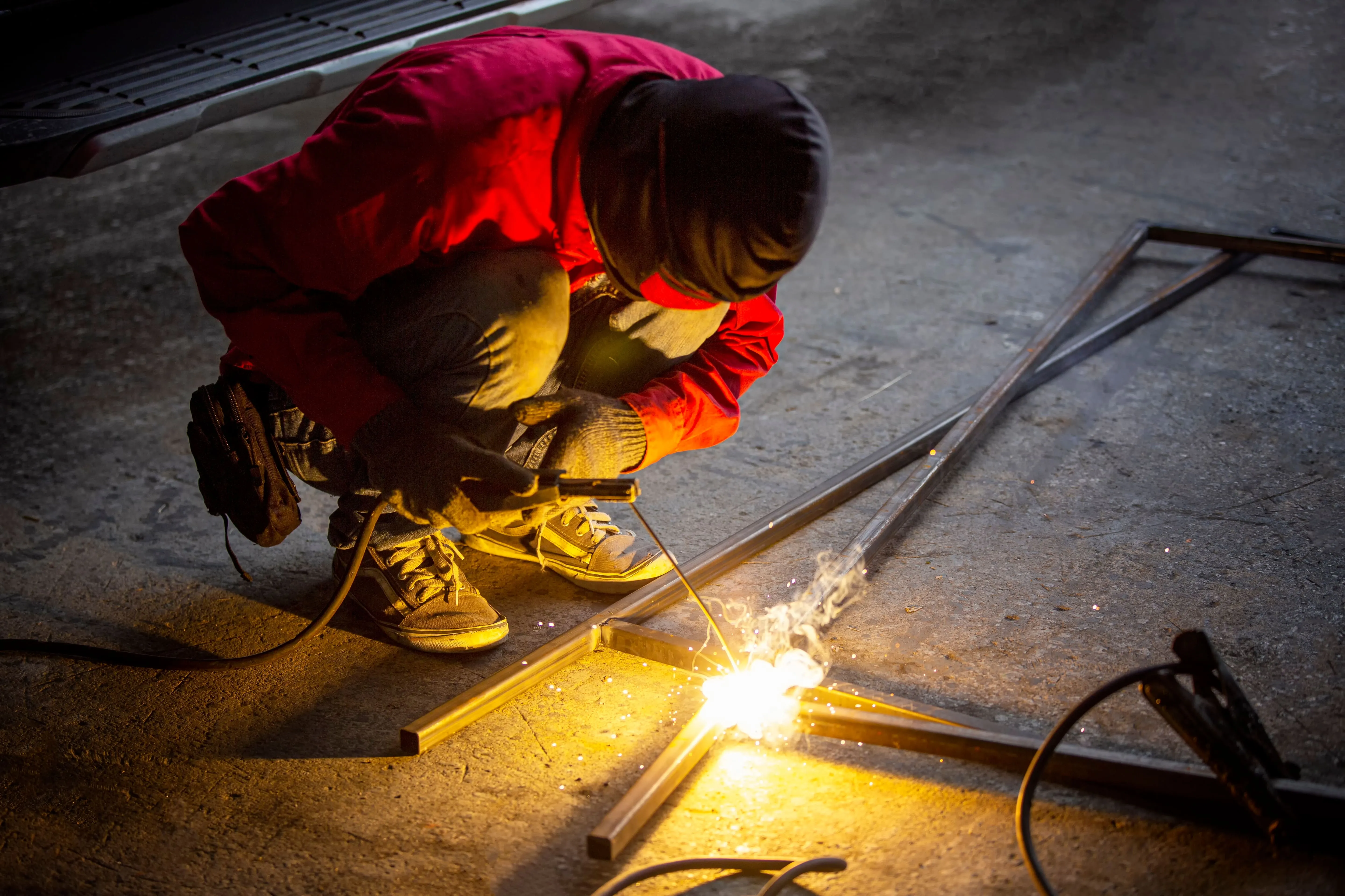 Worker wearing red jacket and gloves welding a steel frame on a concrete floor with sparks.