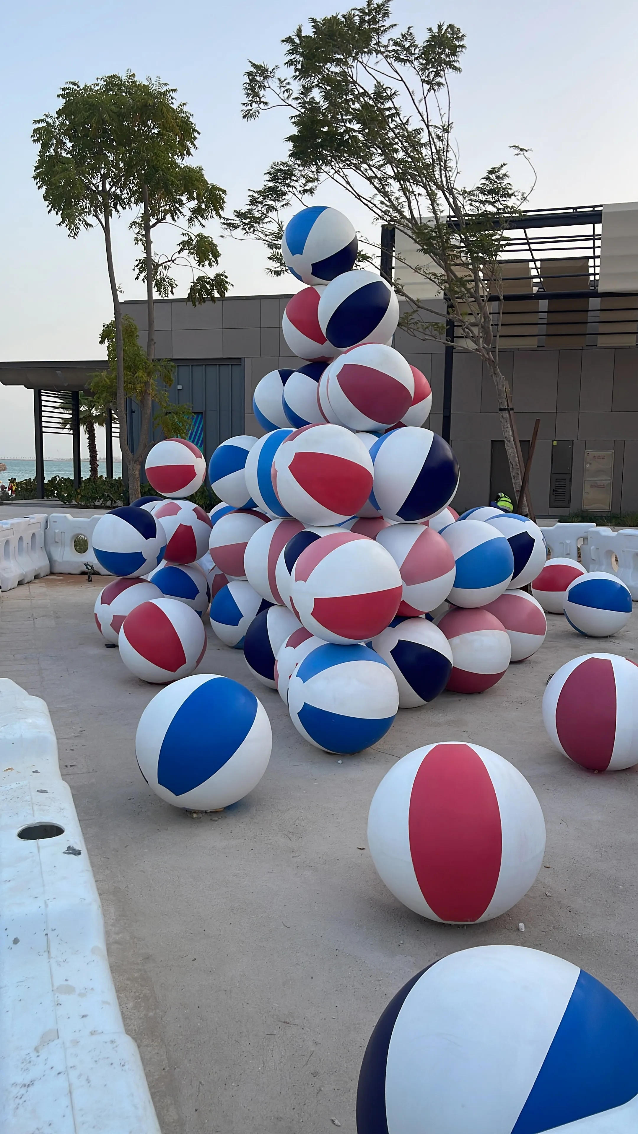 Large pile of red, blue, and pink striped beach balls stacked outdoors near trees and buildings.