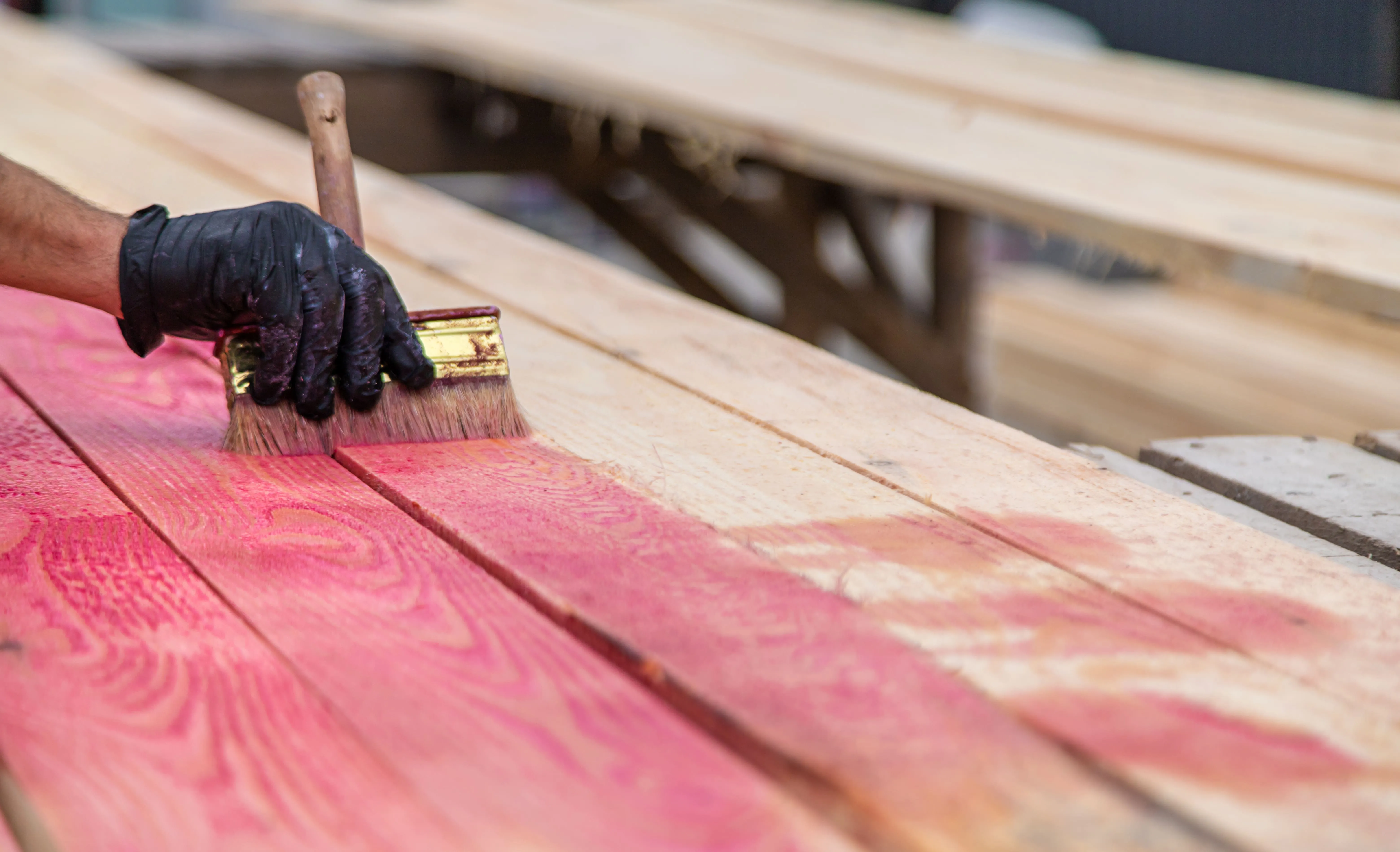 Person wearing a black glove applying red stain or antiseptic on wooden planks with a paintbrush.