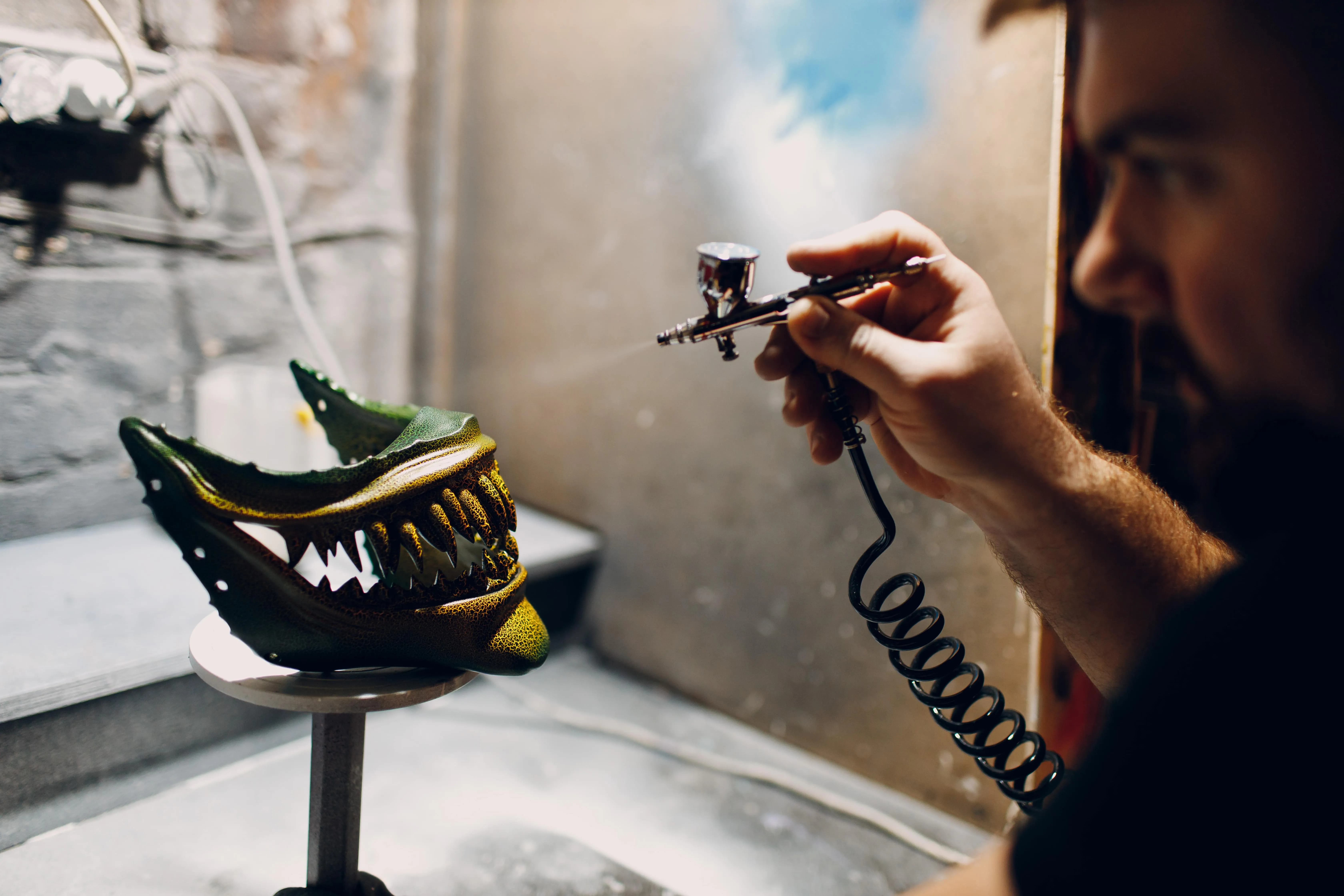 Person airbrushing a green monster mask with sharp teeth on a stand in a workshop.