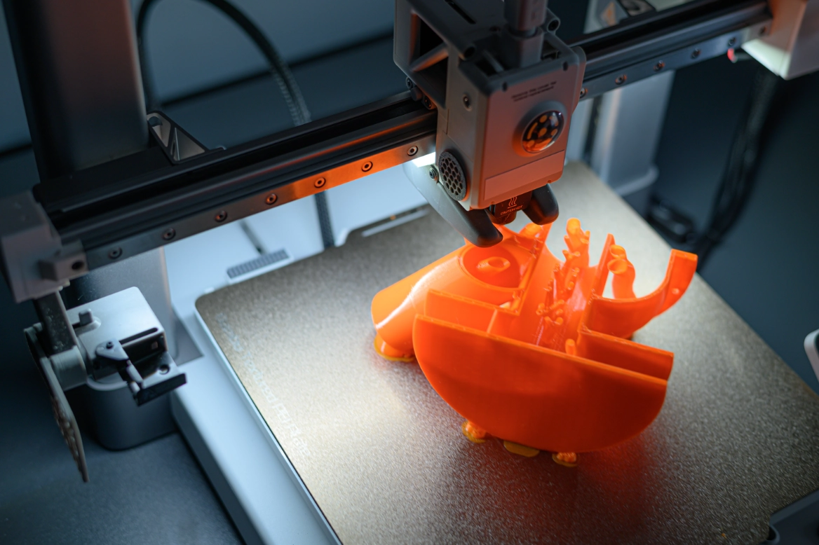3D printer creating an orange plastic object with complex shapes on a print bed.