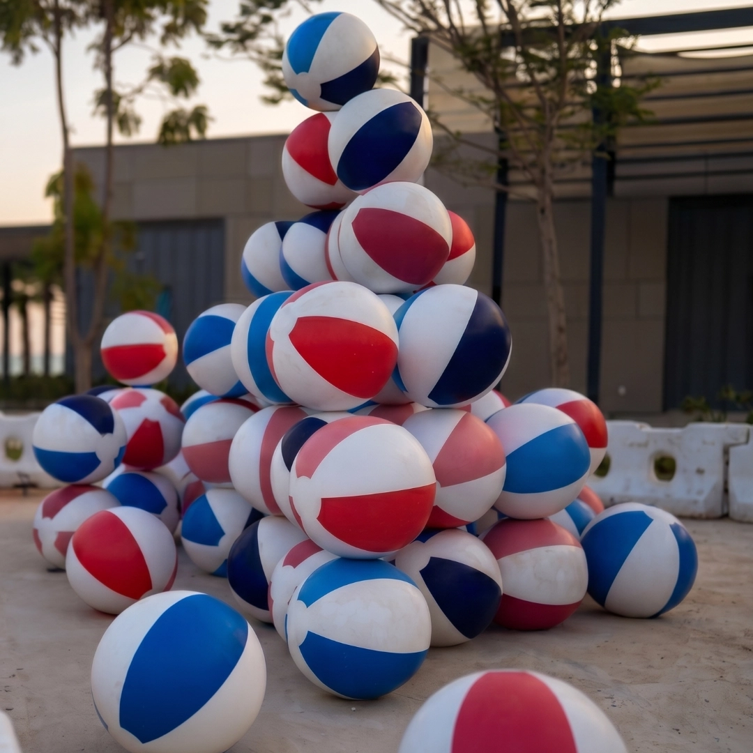 Pile of red, white, and blue beach balls stacked outdoors on a sandy surface near buildings and trees.
