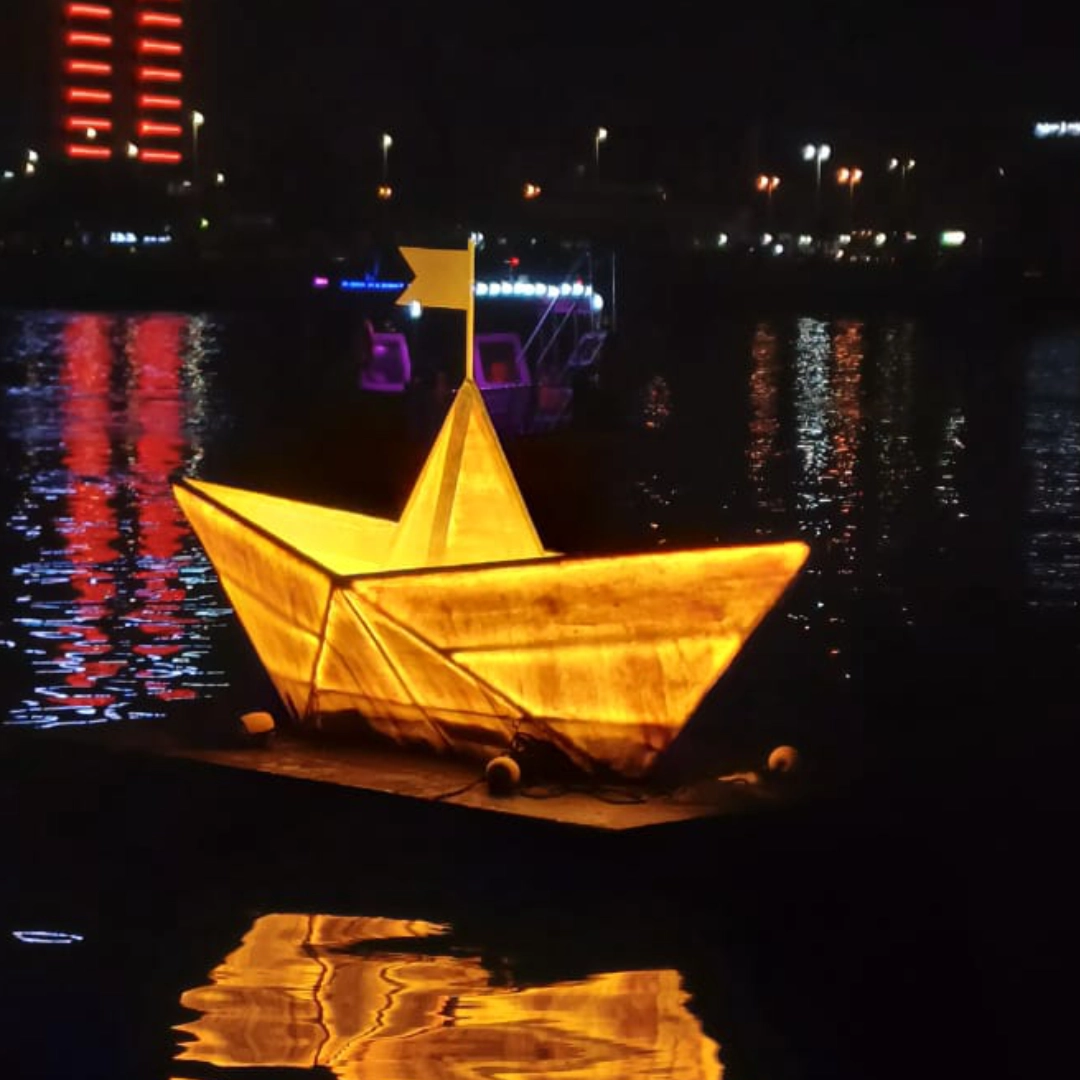 Illuminated yellow paper boat with flag reflecting on water at night near city lights.