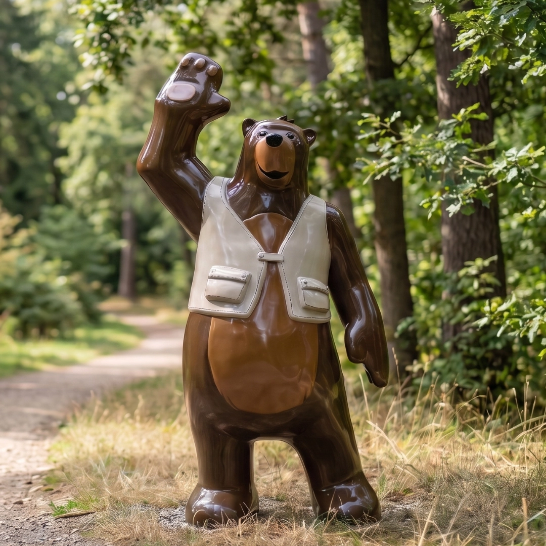 Statue of a friendly bear wearing a white vest waving, standing on a forest path surrounded by trees.