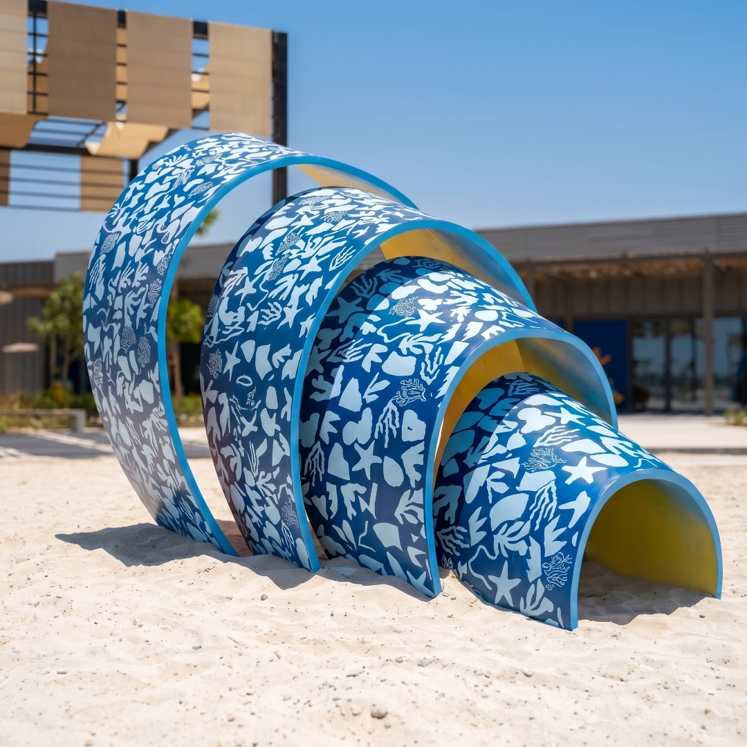 Blue and white sea-themed curved sculpture on sandy ground near a modern building under clear sky.