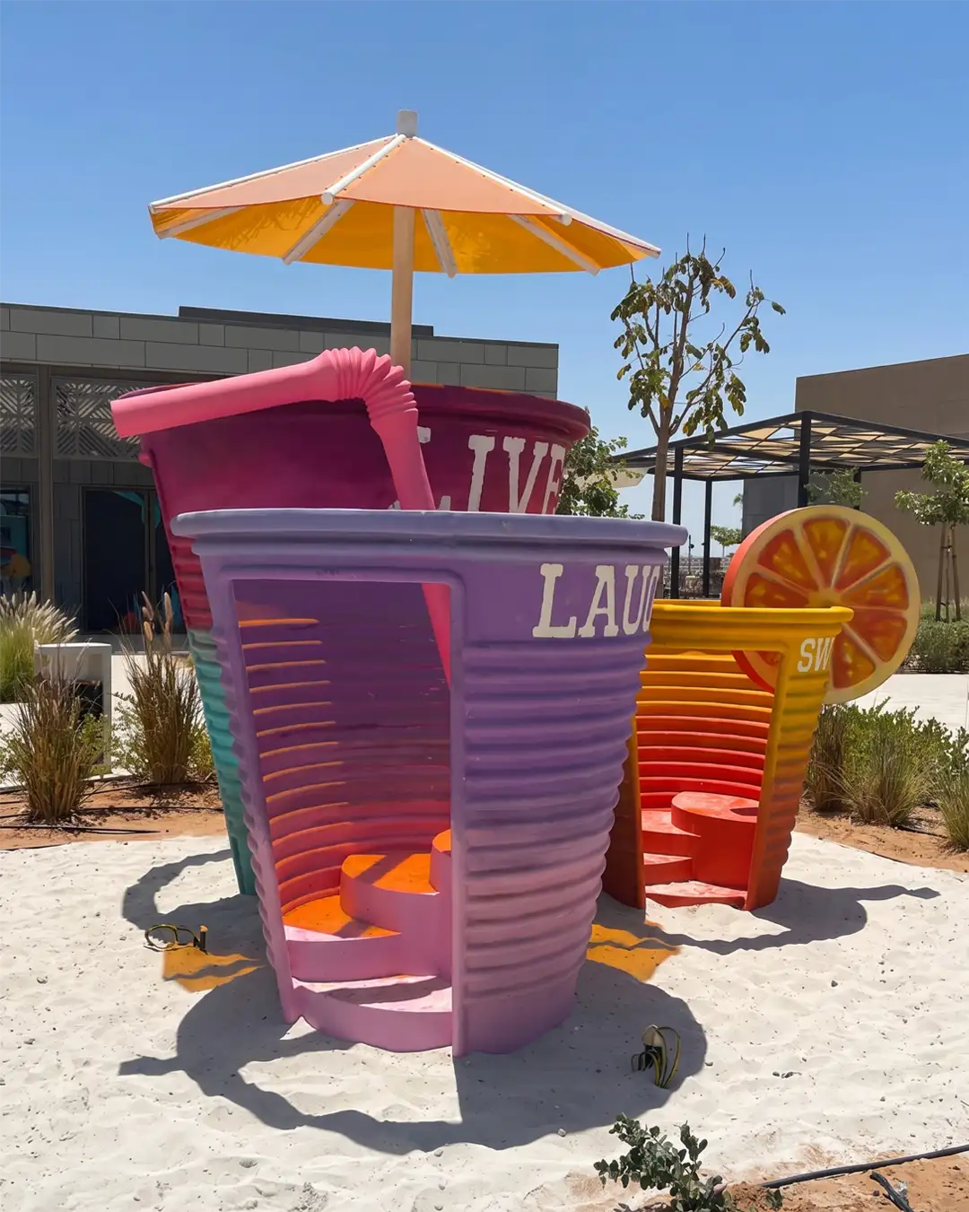 Three colorful oversized cup sculptures with stair entrances in a sunny outdoor area with sand.