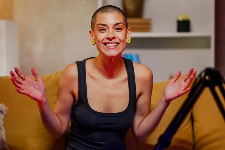 Smiling person with shaved head gesturing with open hands on yellow couch