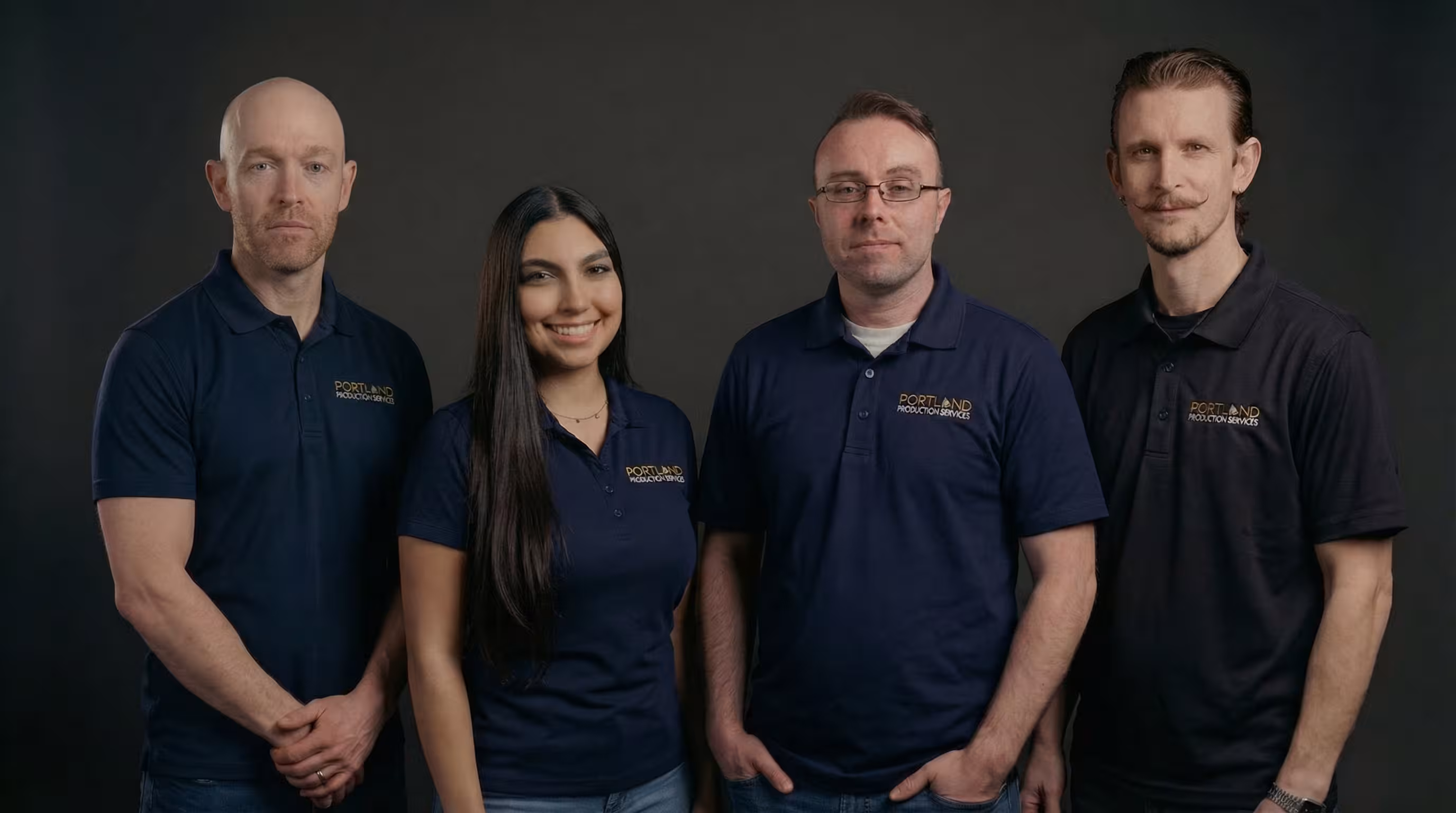 Four people wearing navy blue Portland Production Services polos pose against a dark gray background.
