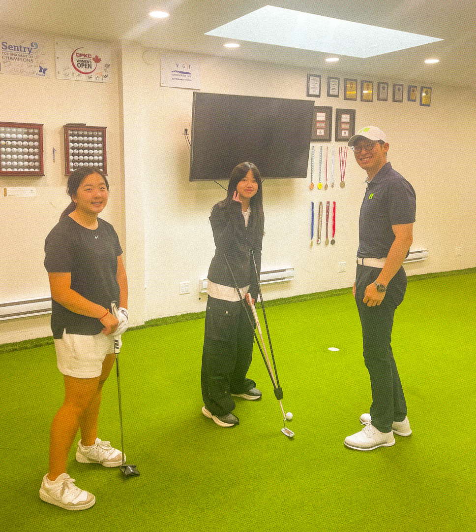 golf instructor smiling next to two students in an indoor golf facility. There is a TV, awards, and golf balls displayed on the wall behind them.