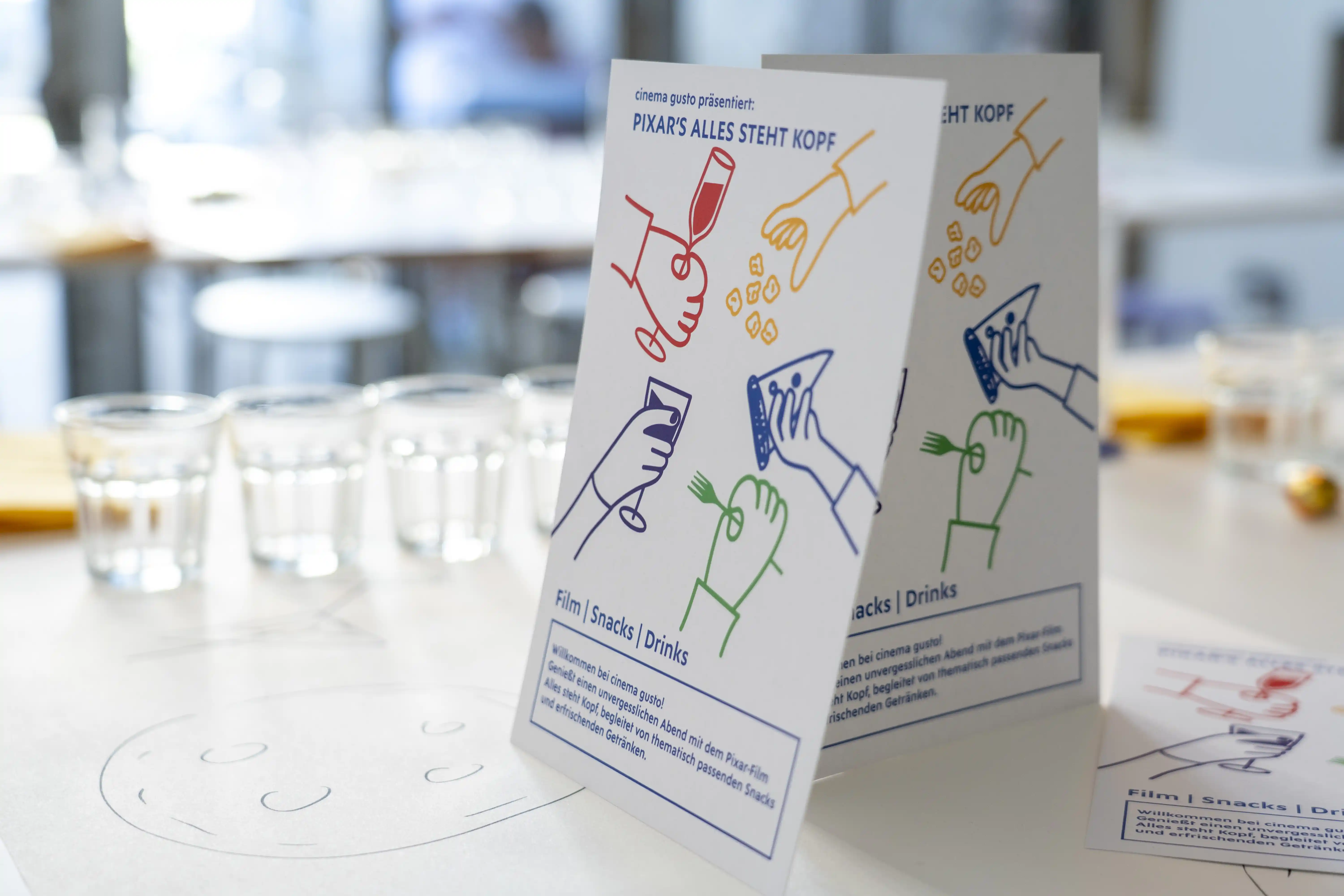 Table tent cards promoting a Pixar-themed event with illustrations of hands holding snacks and drinks on a white table.