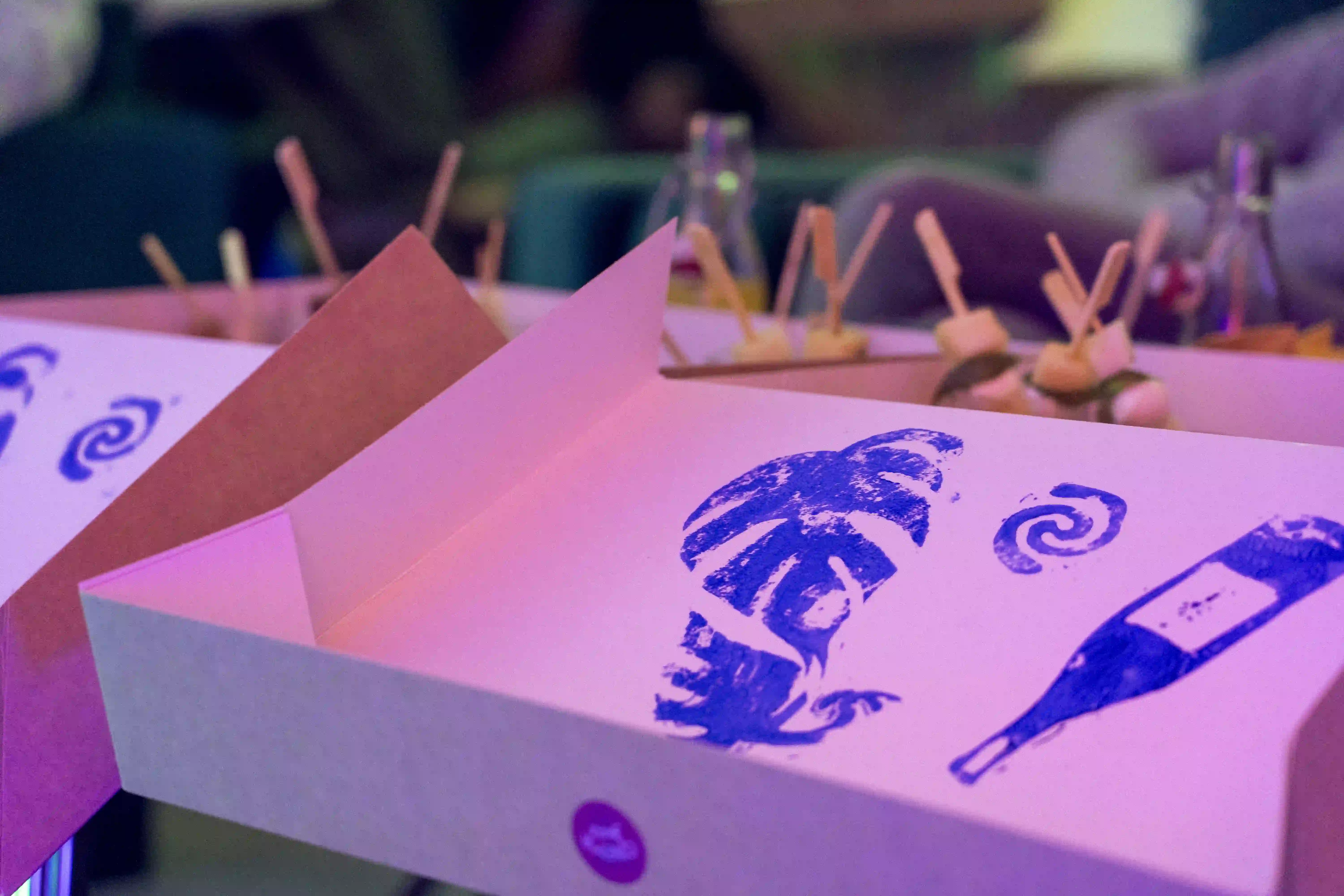 Wine glasses lined up with wooden cones holding popcorn clipped to each glass, set on a table with green glass bottles and a neon pink flamingo light in the background.