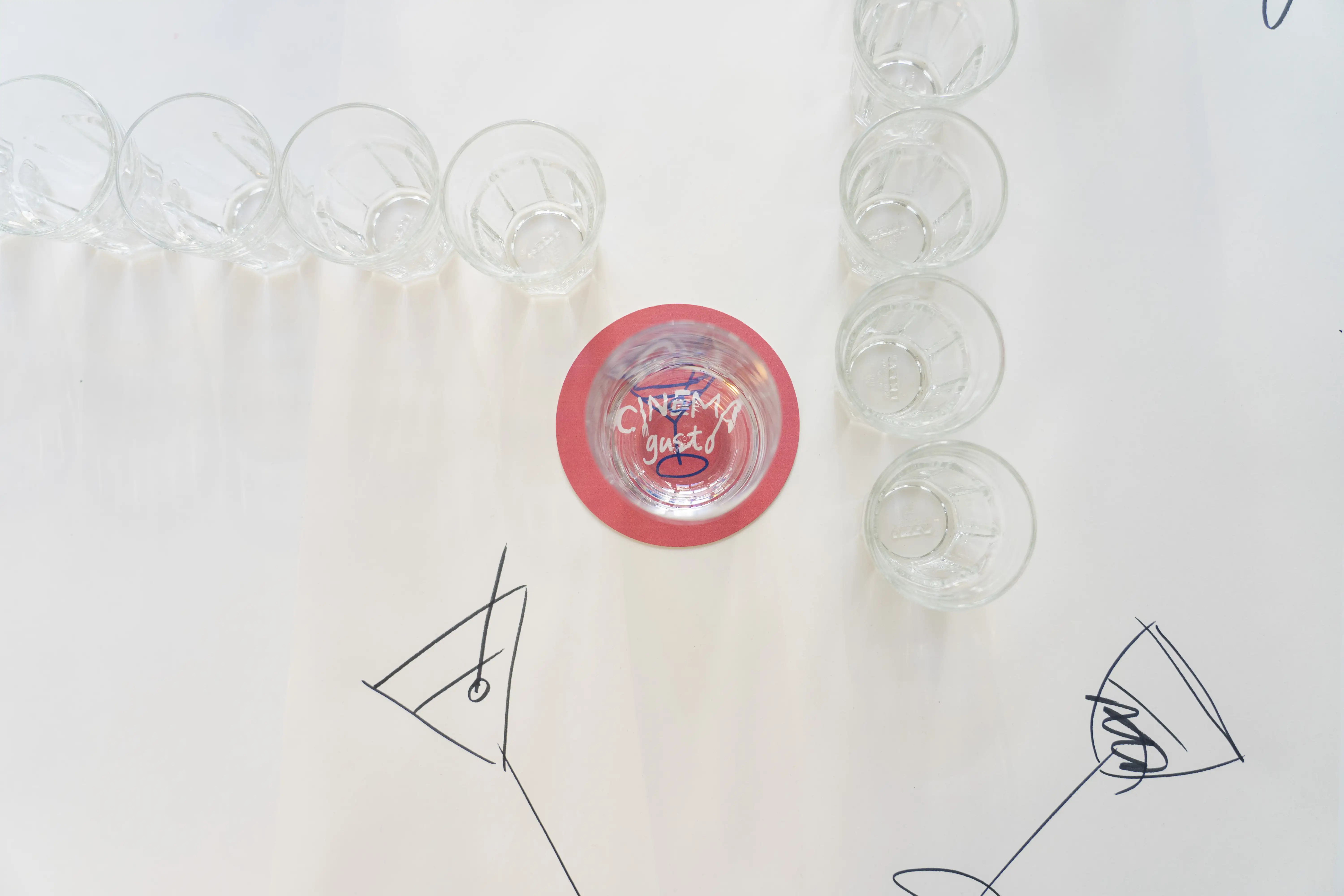 Empty small glasses arranged in lines on a white surface with cocktail glass drawings and a glass on a red coaster.