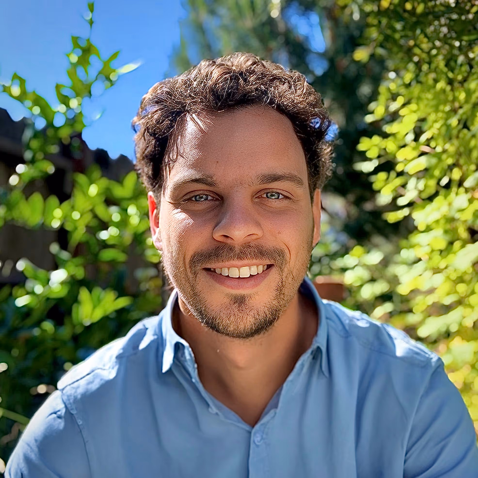 Portrait of Seth Dunn, Architect and Founder of Praxis Architecture, smiling outdoors in a sunlit garden, wearing a light blue shirt.