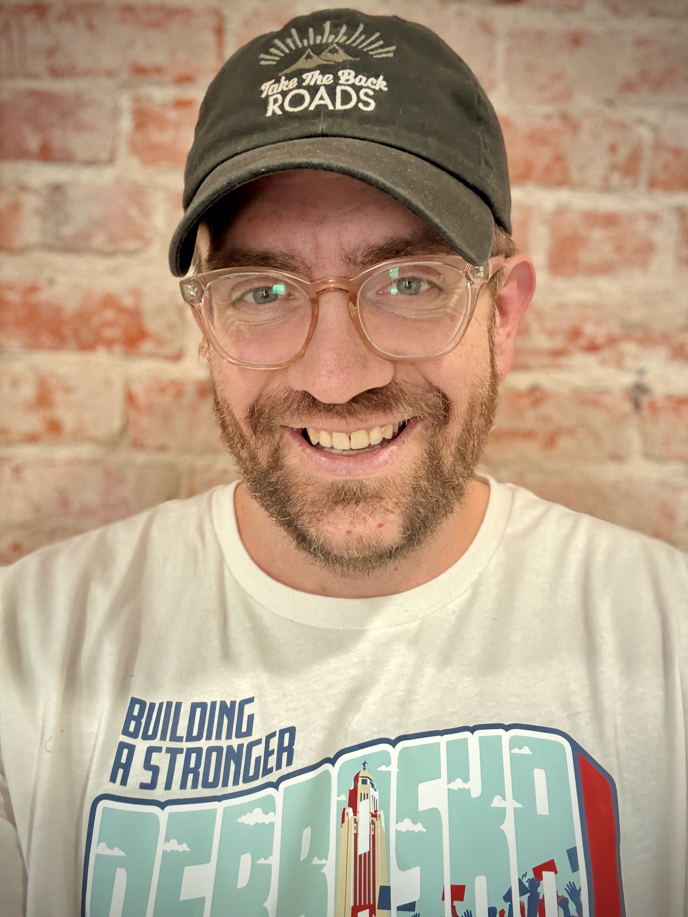 Smiling bearded man wearing clear glasses, a black cap with 'Take The Back Roads' text, and a white t-shirt with 'Building a Stronger Nebraska' text and graphic.