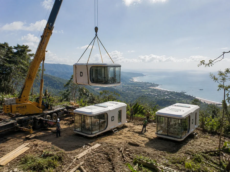 A crane lowering a capsule home onto a hillside site, two units already placed
