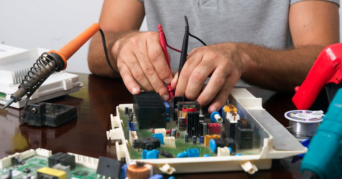 Man working on an electronic circuit board with tools on a table, showing close-up of hands and components.
