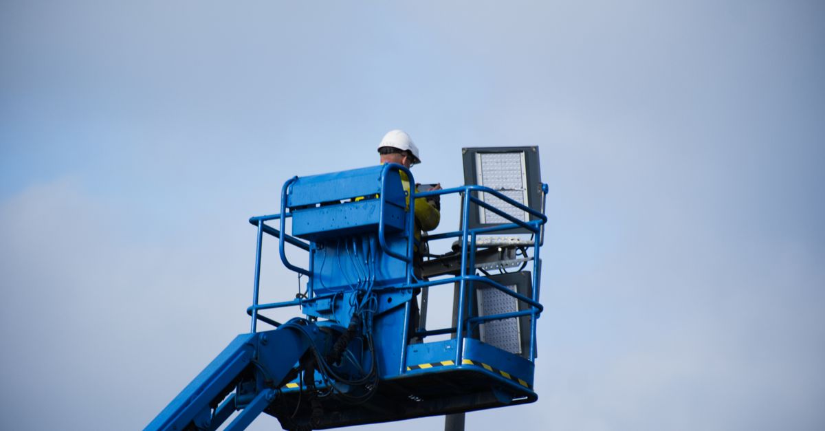 An industrial worker wearing a hard hat operating a lift platform for maintenance tasks in an outdoor setting.