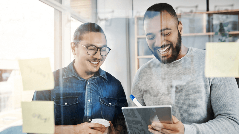 an Asian man & a mixed race man in front of a whiteboard with an iPad