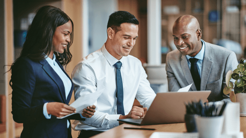 A black woman, a Hispanic man, and a black man, looking through documents and at a laptop