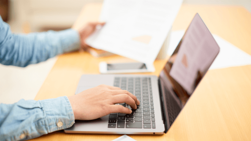 laptop on yellow wood table, white hands in chambray shirt