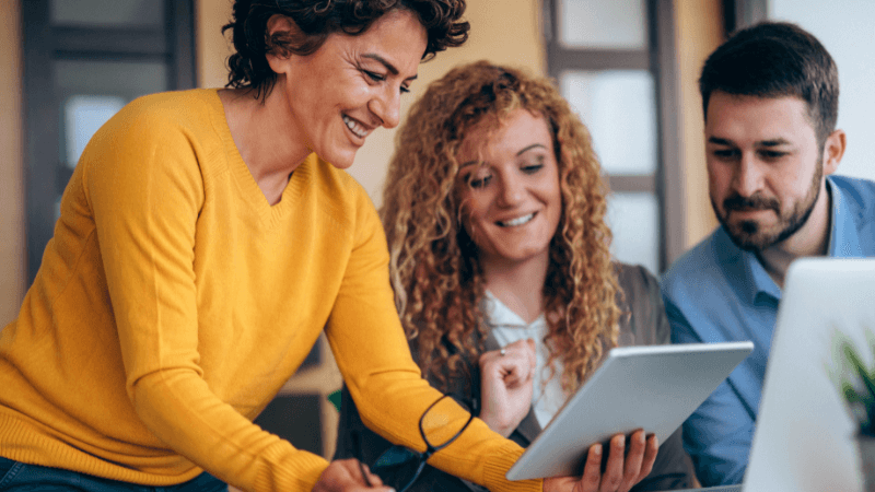 two women, one man looking at iPad and laptop discussing