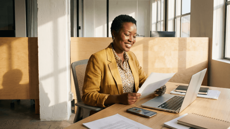a black woman sitting at a desk looking at documents with a laptop