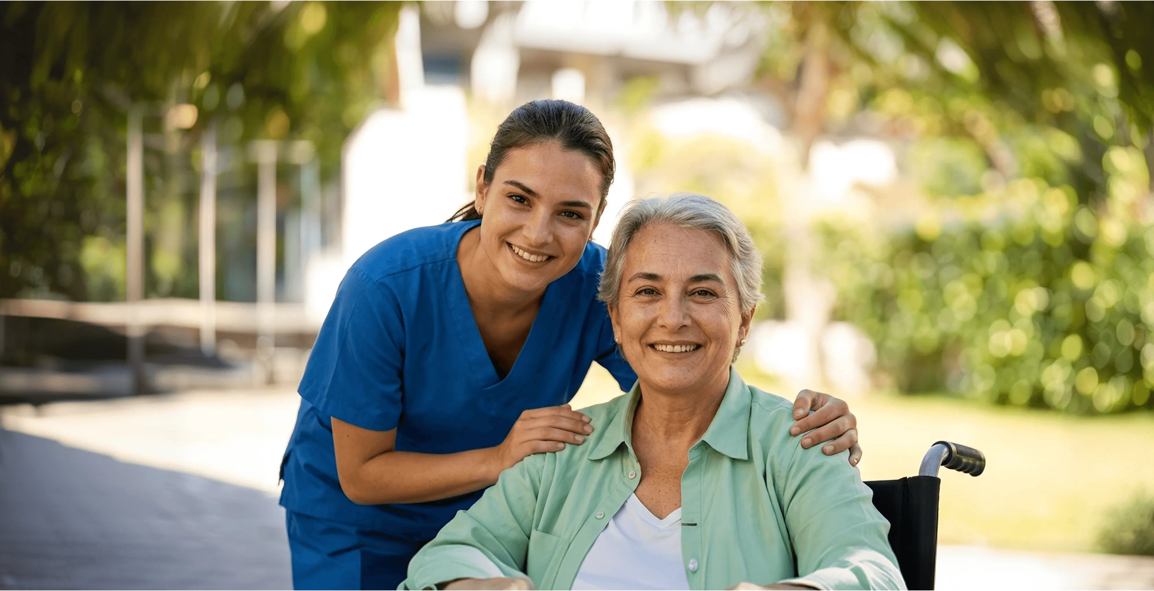 Healthcare worker in blue scrubs standing beside smiling elderly woman outdoors
