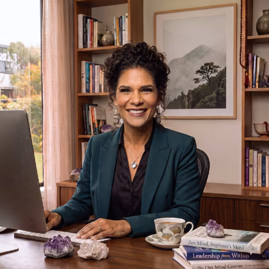 A smiling woman with dark, coily hair styled in an updo sits at a warm wooden desk. She is wearing a teal blazer over a black button-down shirt, a small nose stud, tiered earrings, and a silver heart pendant necklace. Her hands rest on a white computer keyboard next to a large monitor. The desk is decorated with raw amethyst and clear quartz crystals, a teacup featuring a ginkgo leaf design, and a stack of books including Zen Mind, Beginner's Mind and Leadership from Within. Behind her is a wooden bookcase filled with books and decor, and a framed picture of a misty mountain landscape hangs on the wall. A window with a view of outdoor greenery is visible to the left.