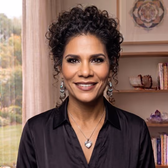 A headshot of a smiling woman looking directly at the camera. She has dark, curly hair styled in an updo with loose tendrils framing her face. She is wearing a black button-down collared shirt, long tiered earrings with turquoise accents, a small nose stud, and a silver heart pendant necklace. Behind her is a soft-focus background featuring a window with sheer curtains on the left, and wooden shelves on the right holding books, a small bowl, crystals, and framed geometric artwork."