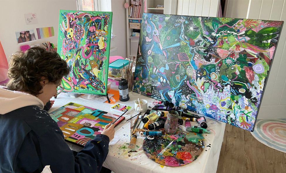 Artist Sian Atkins sits at a desk, painting a colorful abstract piece. Two finished canvases are propped up nearby. The workspace is lively, with brushes, paint tubes, and a palette scattered across the table.