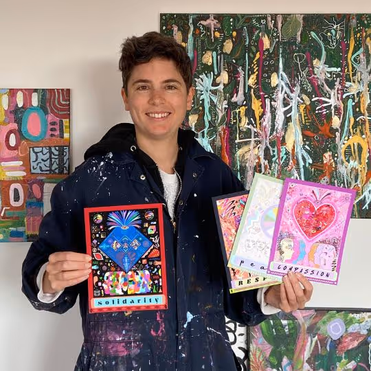 Sian Atkins is holding four illustrated art prints labeled “Solidarity,” “Peace,” “Respect,” and “Compassion” in a studio with paintings behind her.