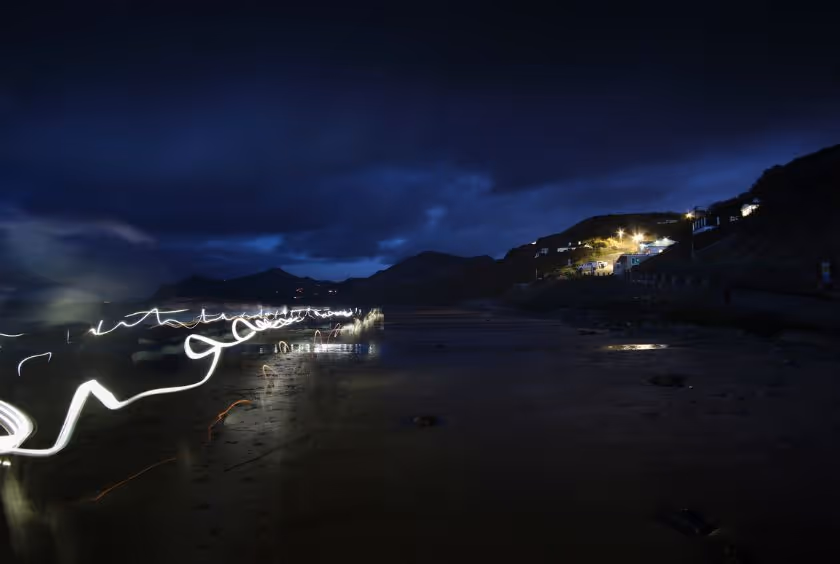 Art work by Giles W. Bennett. Long exposure photograph in the evening, on a beach. Mountains in the background and lines of colored lights on the left.