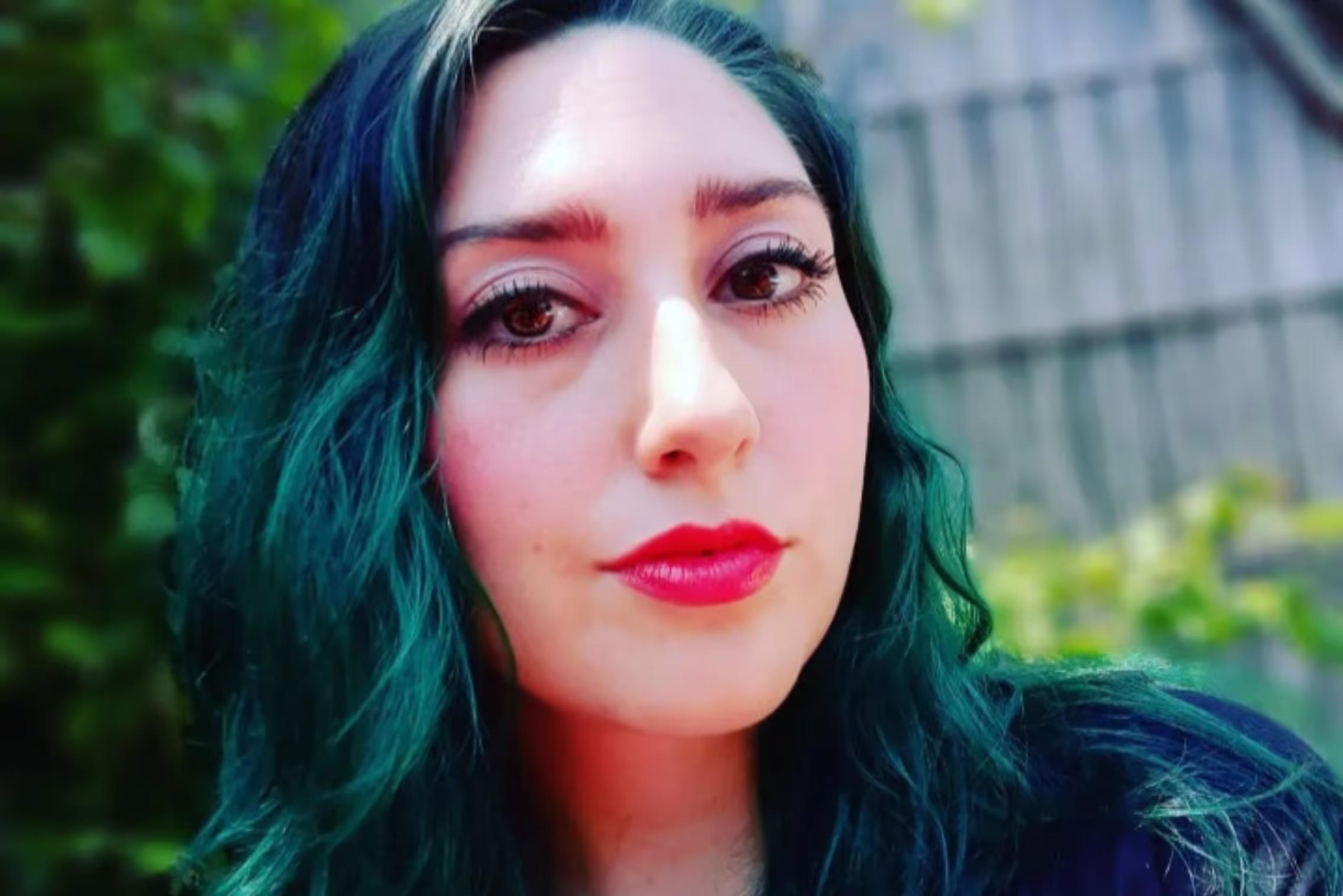 A woman with long, wavy teal-colored hair poses outdoors, wearing a dark V-neck shirt. She has a confident expression with bright red lipstick, and the background features lush green foliage and the blurred outline of a wooden structure.