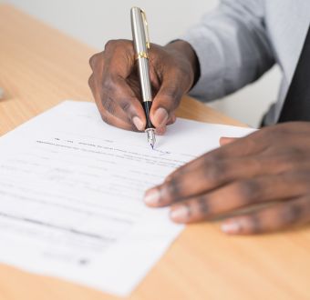 Hands on a sheet of paper. One of the hands holds a pen and is signing the document.