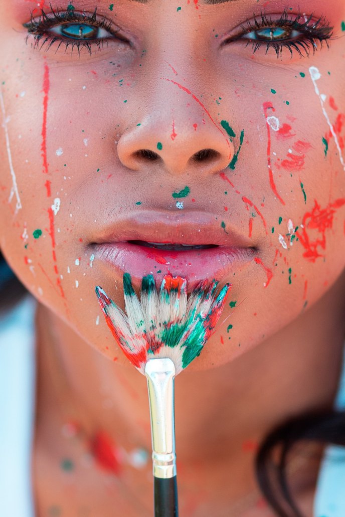 An attractive BIPOC female energy artist with paint on their face. There is a fan paintbrush on their chin.