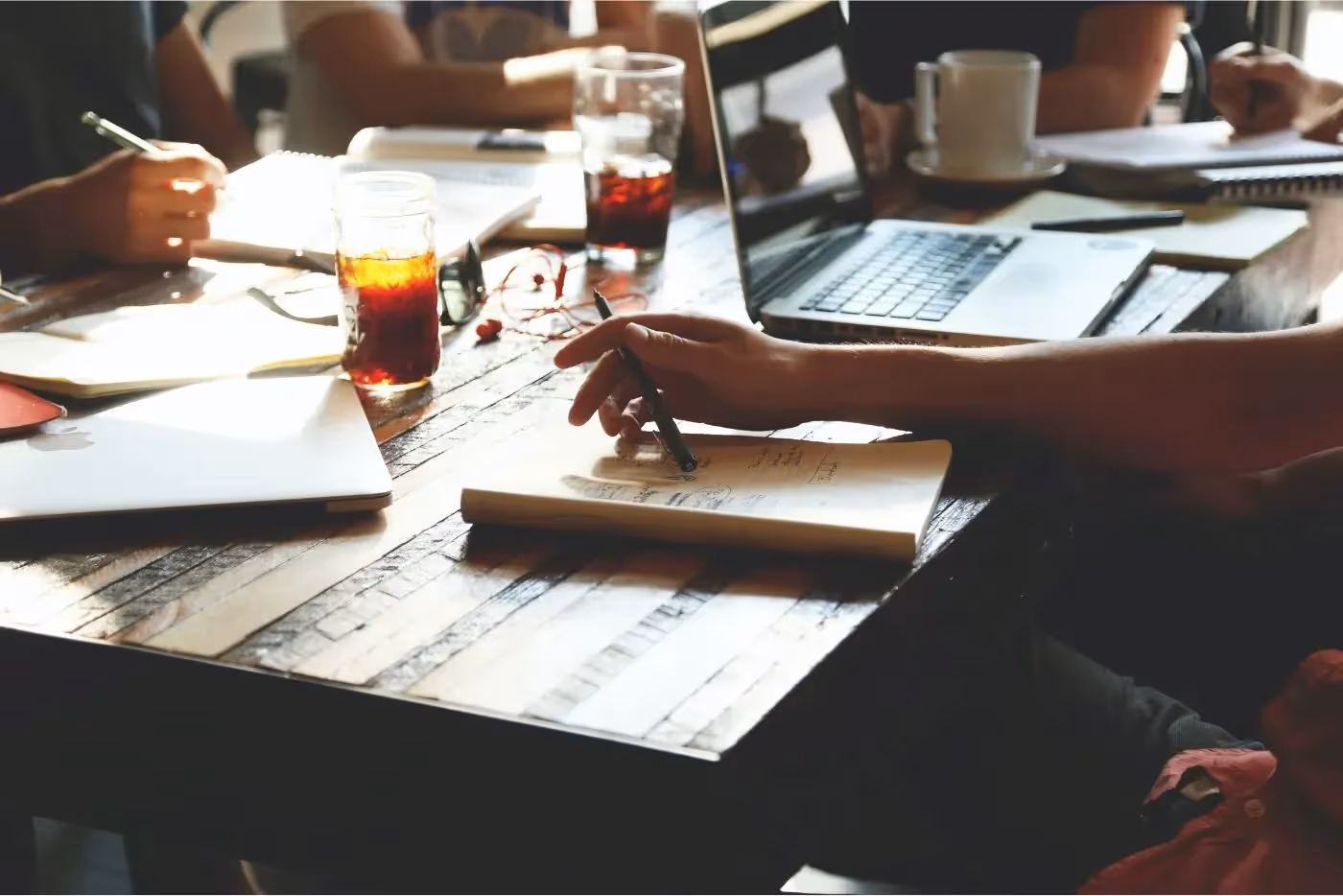 Photo of a sunlit desk. There are notepads, laptops, cups of beverages, and the hands of four people sitting there, you cannot see their faces, only partial torso and hands holding writing tools like pencils and pens.