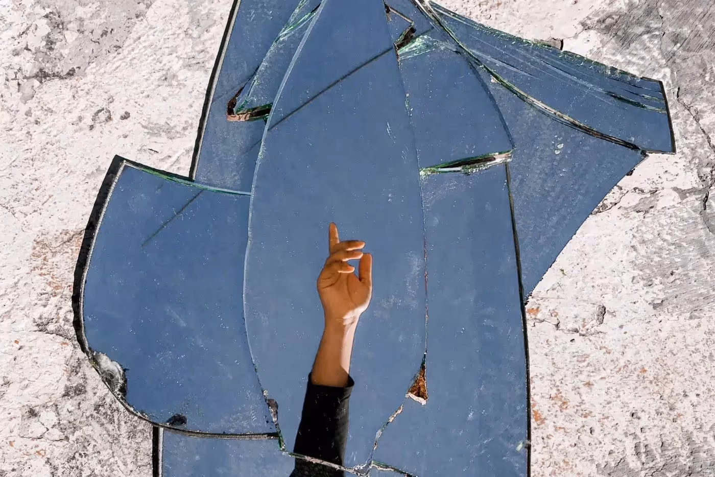 Photo of a broken mirror with a reflection of an arm raised wearing a long sleeve dark shirt. It symbolizes, "I'm here". Photo by Bruno Pires from Pexels: https://www.pexels.com/photo/mirror-fragments-on-gray-surface-with-the-reflection-of-a-person-s-hand-5514777/