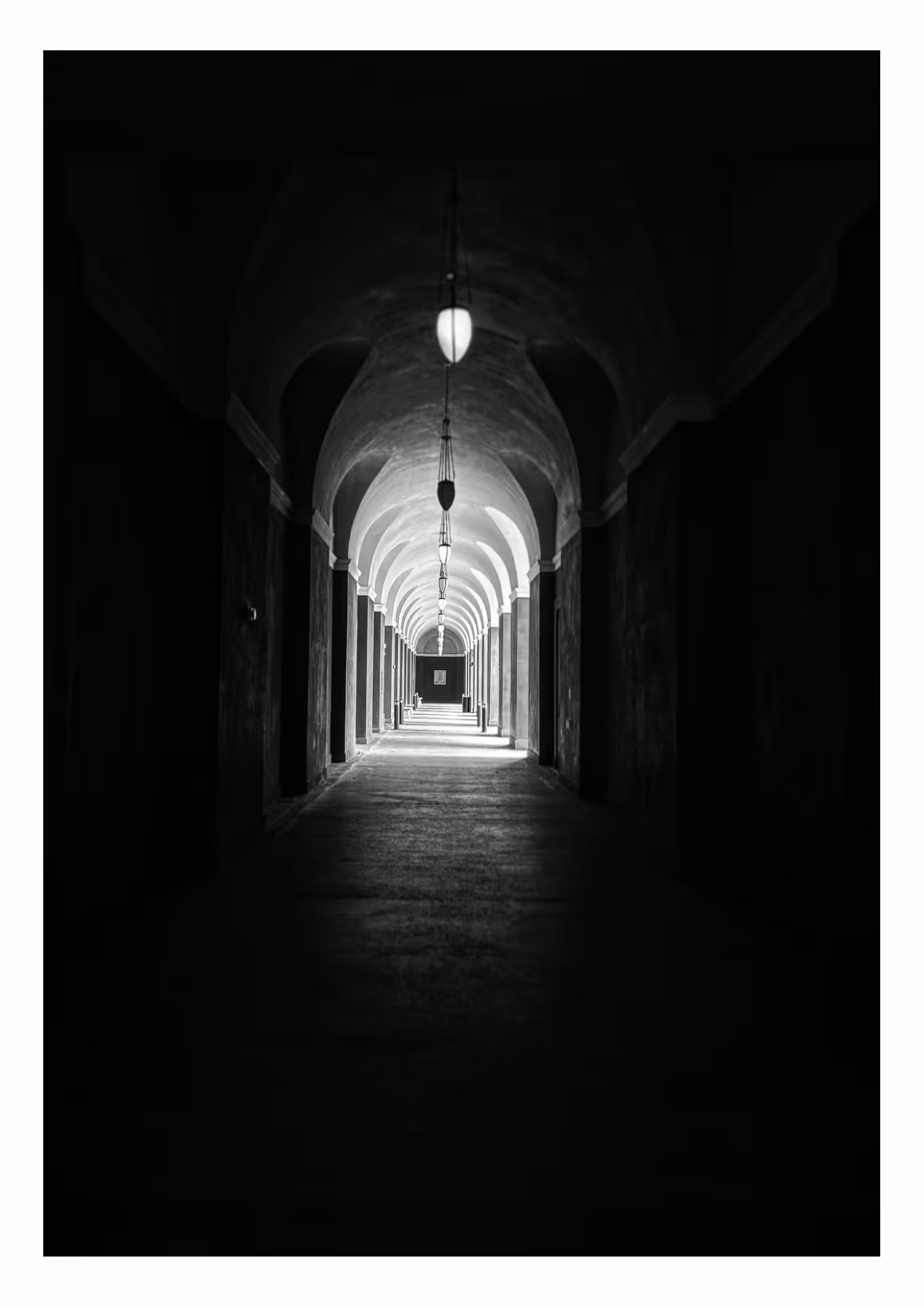 Black and white photo of a long hallway with very high rounded cathedral like ceilings. There are lamps hanging from the ceiling on chains.