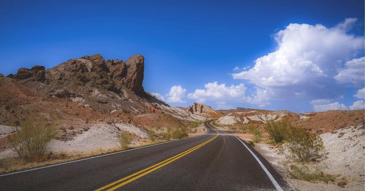 A winding two-lane highway cuts through the rocky desert landscape of West Texas, with rugged hills on each side and a bright blue sky filled with large white clouds overhead.