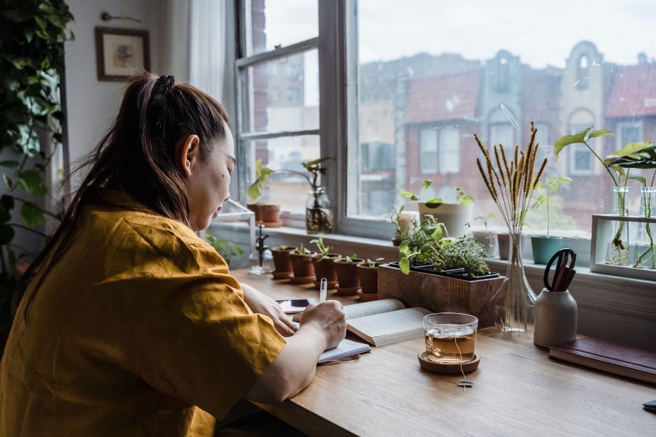 A woman wearing a yellow top sits at a wooden desk by a window, writing in a notebook next to a glass mug of tea and a row of small potted green plants.