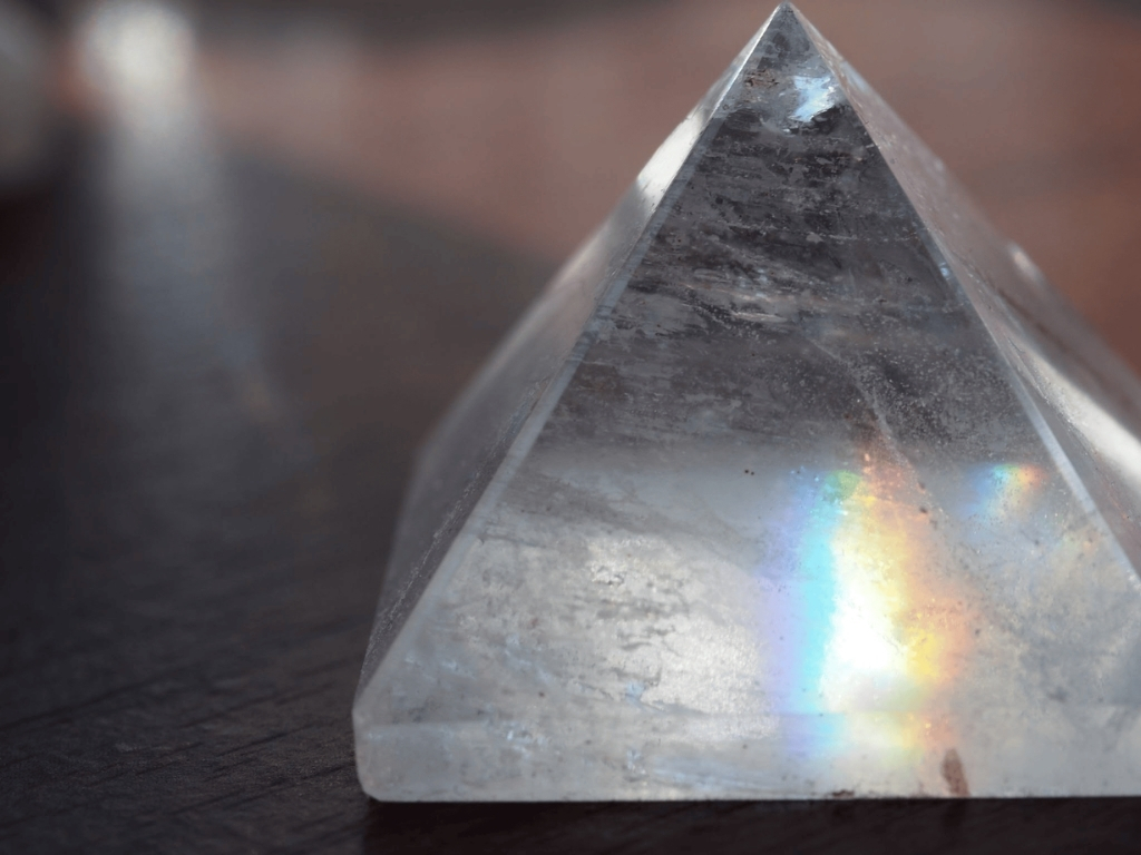 A clear quartz crystal pyramid sitting on a dark wooden surface, reflecting a bright rainbow prism of light on its lower right side.