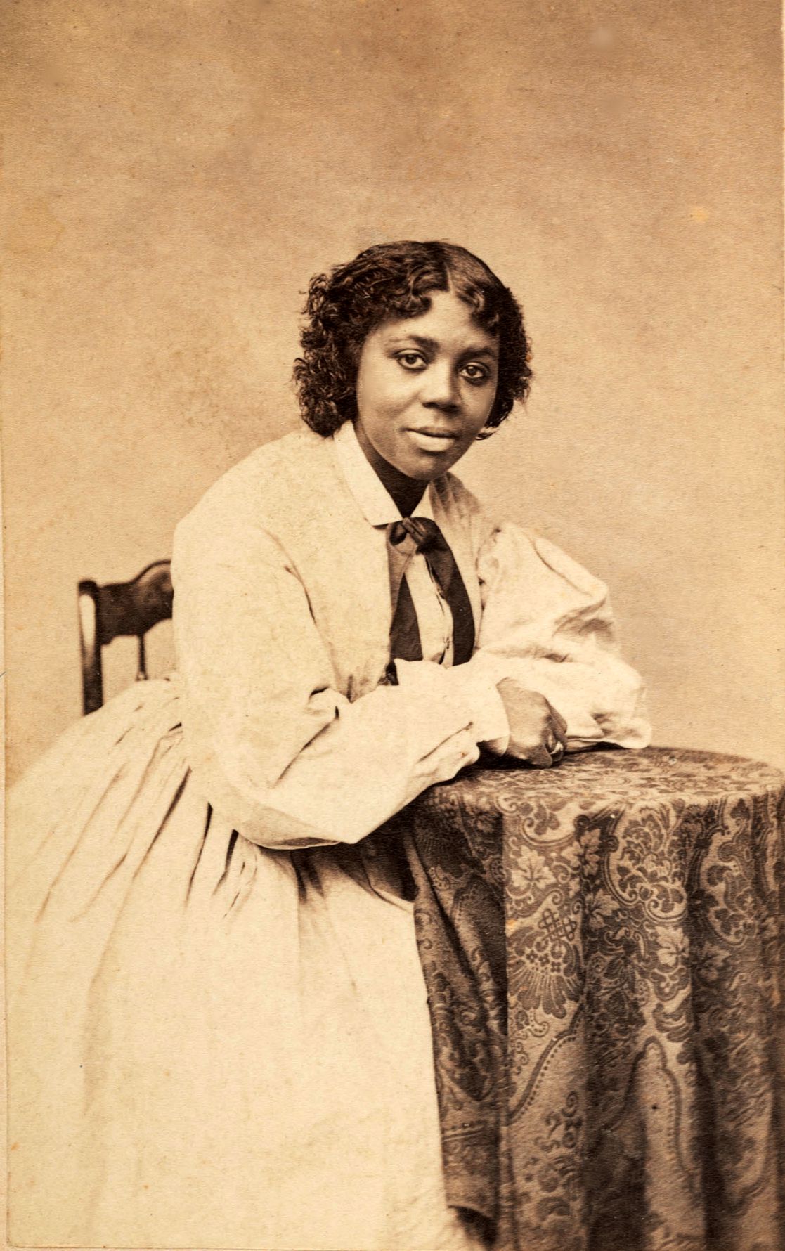 Sepia photograph of Edmonia Lewis around 1870, seated with her arms resting on a patterned tablecloth, wearing a light dress with a dark ribbon at her collar.