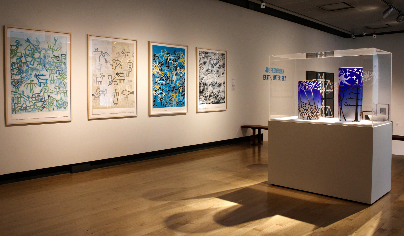 A gallery with bright, colorful prints hung along white walls and a glass case in the center holding blue and multicolored glass vessels.