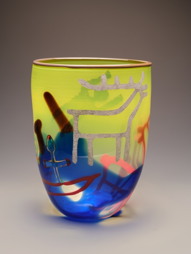 A blown glass vessel that shifts from bright yellow-green at the rim to deep blue at the base, marked with faint white line figures and a brushy red-brown stroke.