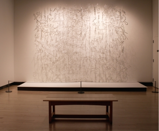 A long gallery wall covered in rows of small, pale hanging glass forms, with a wooden bench in front.