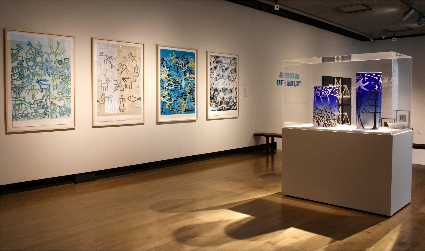 A gallery with bright, colorful prints hung along white walls and a glass case in the center holding blue and multicolored glass vessels.