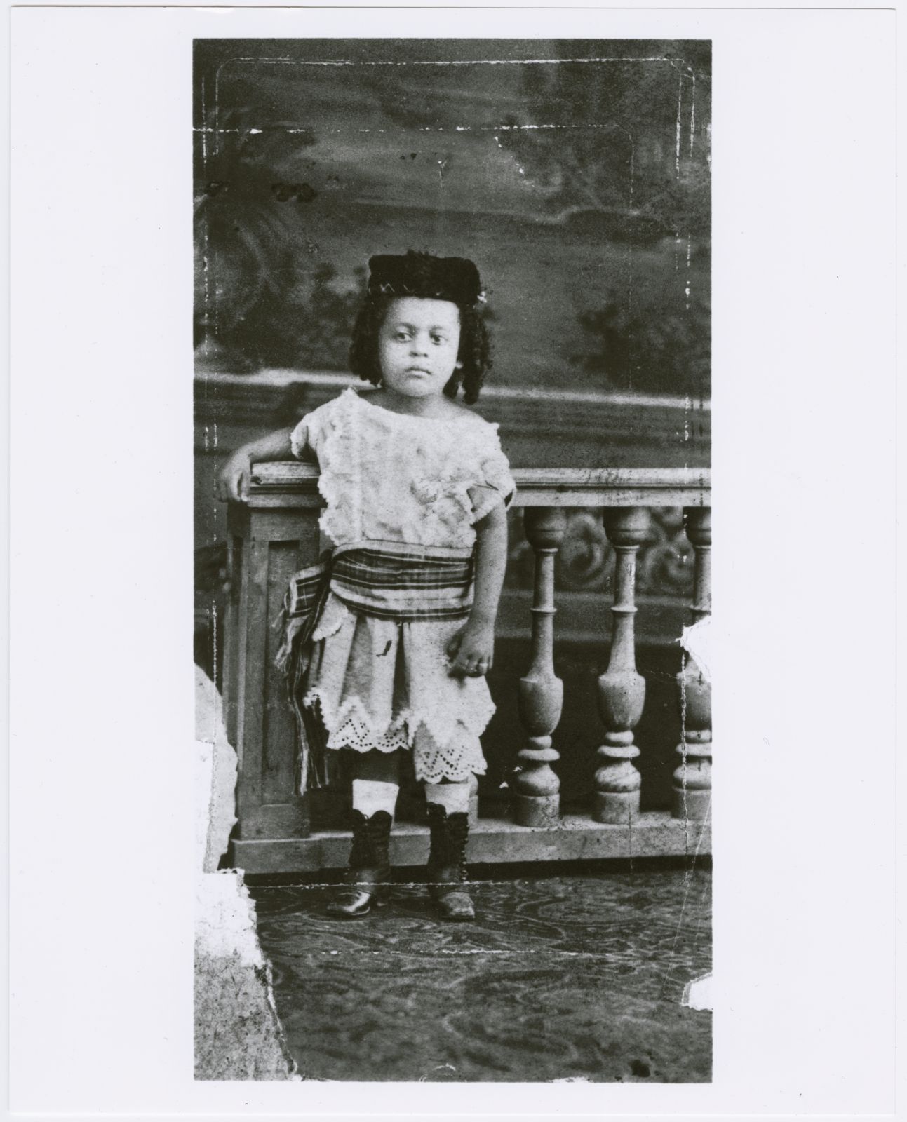 A worn 1878 studio photograph, shown as a print on a pale gallery wall. Four-year-old Schomburg stands beside a turned balustrade in a light dress with eyelet trim, what looks like a plaid sash, a dark cap, and laced boots. The print’s edges are torn.