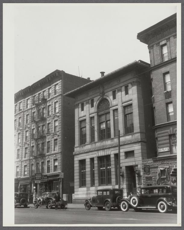 A black-and-white photograph, shown as a print on a pale gallery wall, of a stone library building with tall arched windows on a Harlem street. It stands between brick apartment buildings, with 1930s cars parked at the curb.